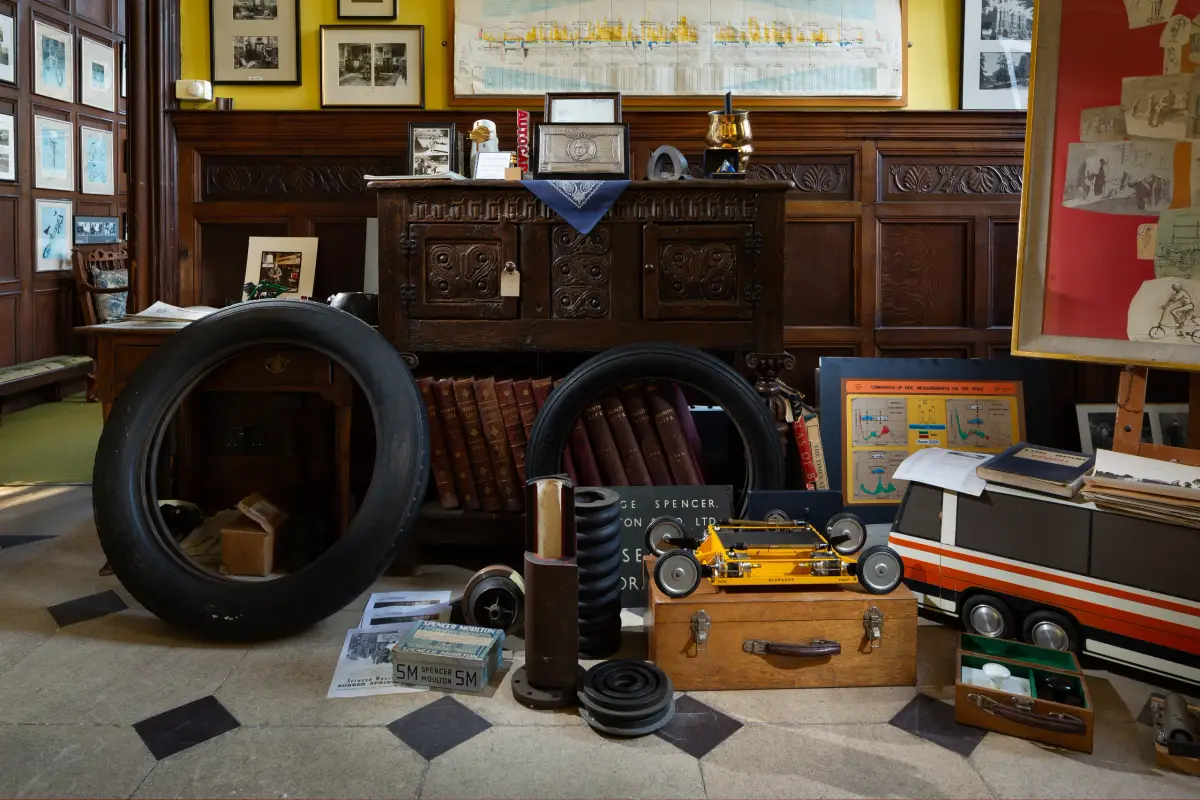 A panelled room hung with framed drawings, a carved oak chest behind a display of bicycle tyres, suspension parts and a model car laid out on the floor