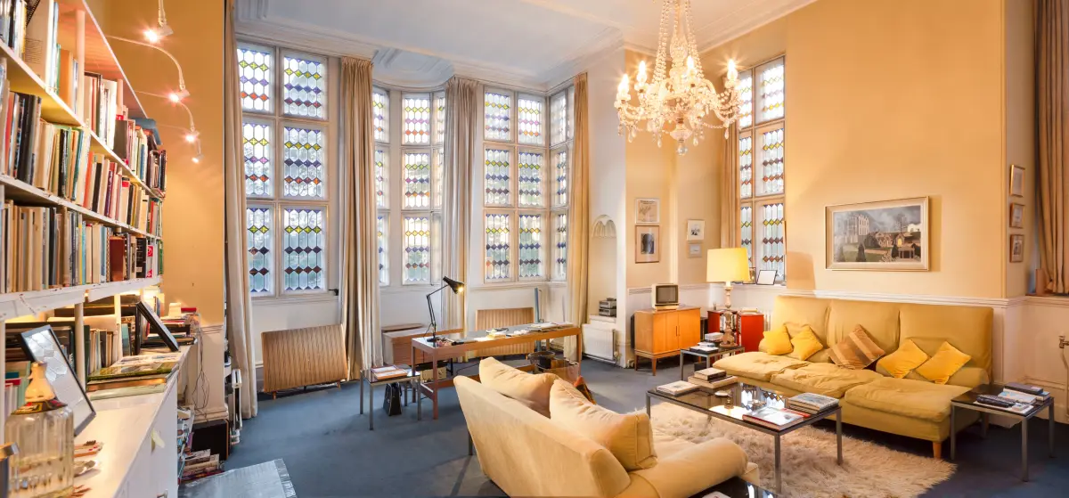 A high yellow drawing room with a chandelier, bookshelves to one side and sofas facing tall stained glass windows