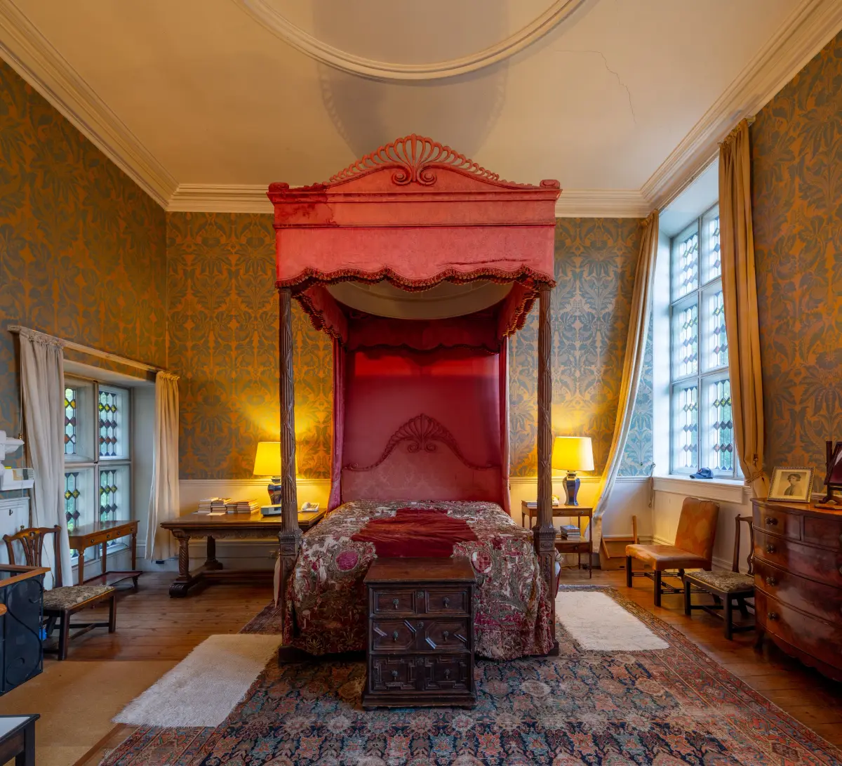 A bedroom with a red canopied four poster bed against patterned blue and gold wallpaper, windows to two sides and a worn rug across the boards