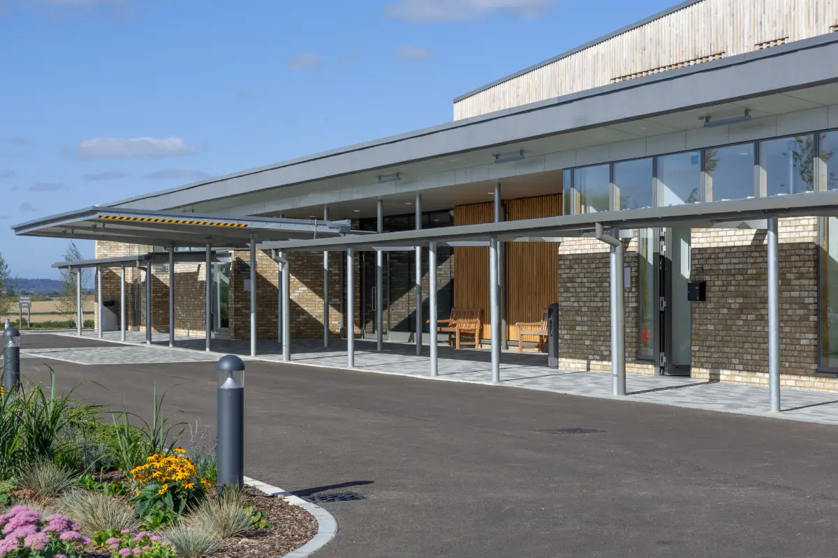 The approach to the building, a slender steel colonnade carrying a deep canopy along a glazed front of brick and vertical timber