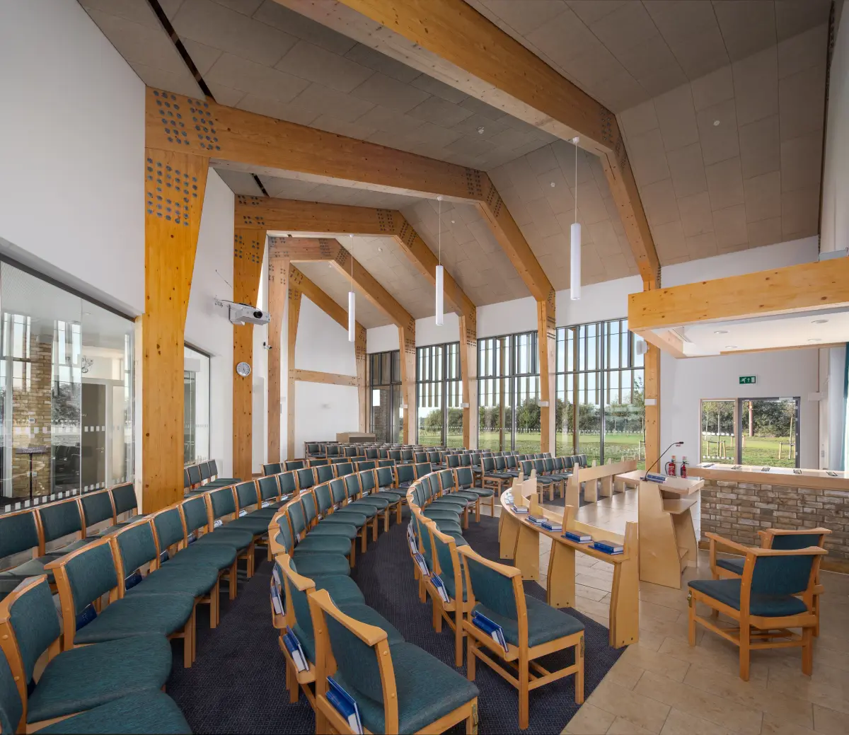 The chapel from the rear, curved rows of seating under exposed timber trusses facing a lectern and a wall of glass onto the fields
