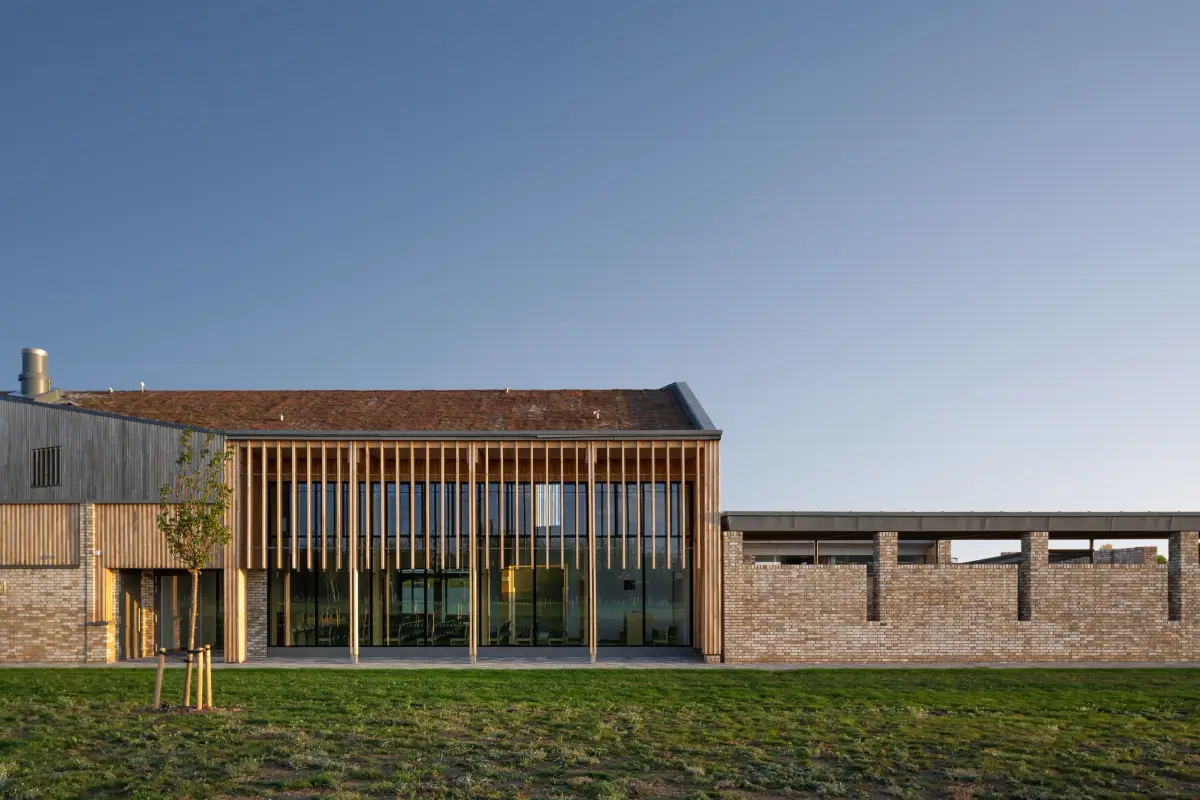 The building from the meadow at low sun, a tiled roof over a tall timber screen and a long brick wall running out to one side
