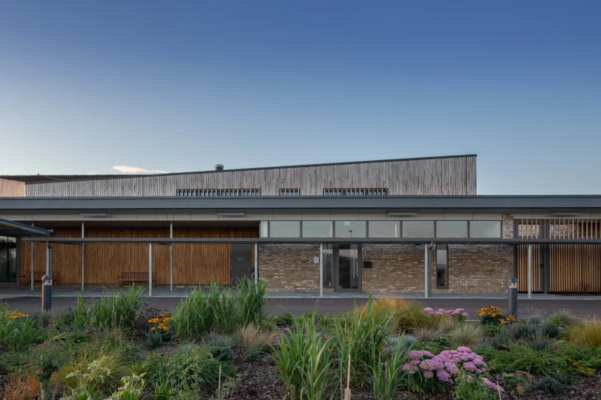 The long elevation at dusk behind a planted border, timber cladding above brick and a covered walk along the front