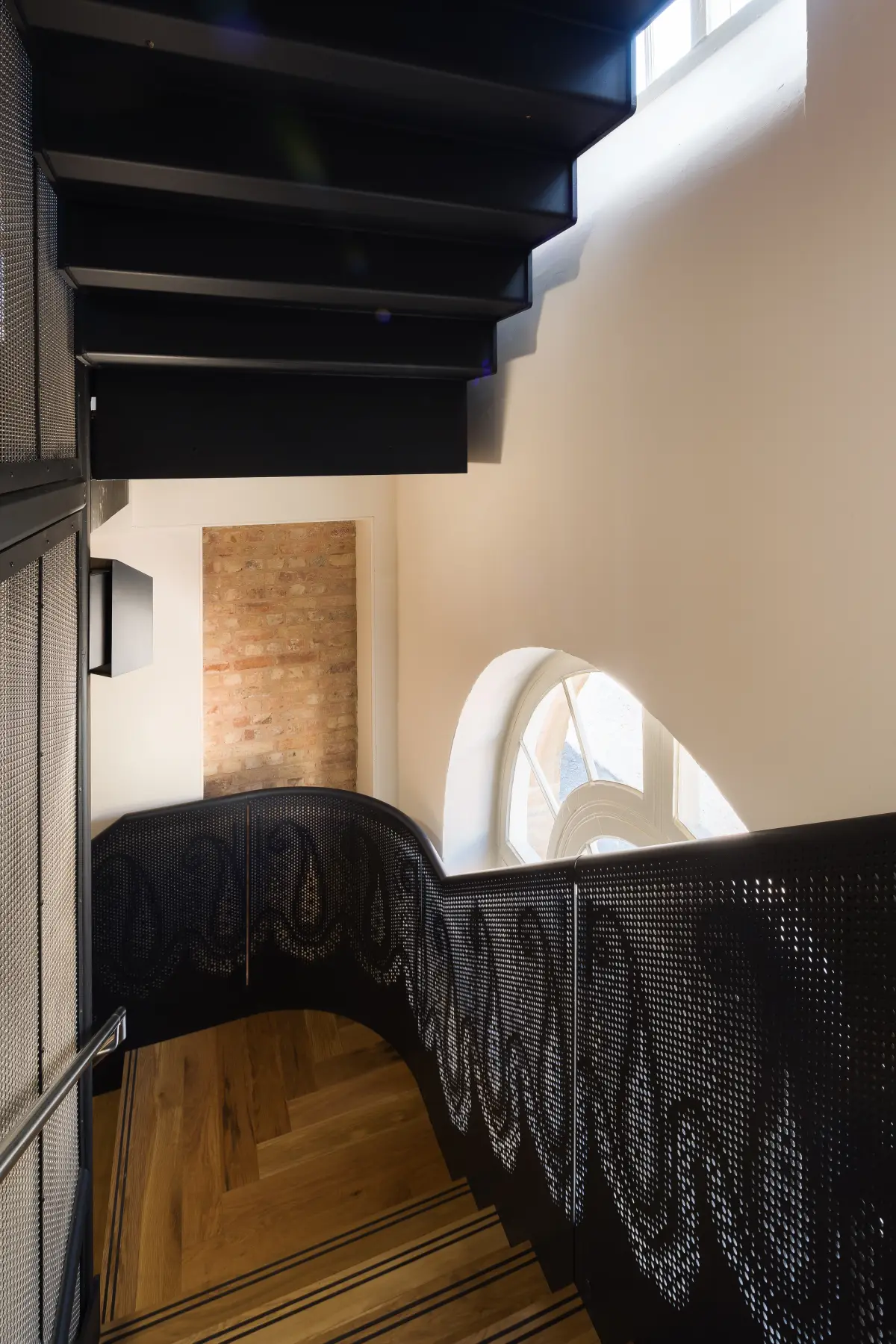 A black steel stair rising past a balustrade cut with a foliage pattern, a round-headed window and a panel of exposed brick beyond