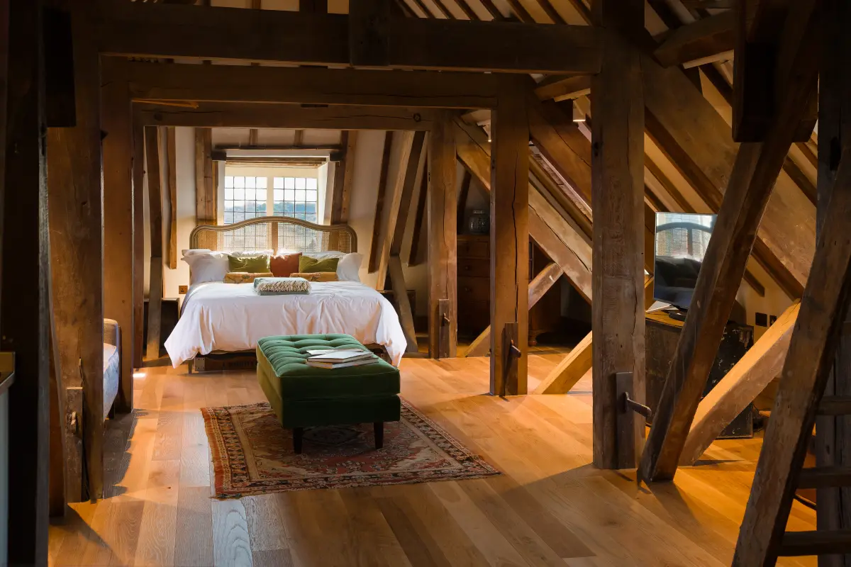 An attic bedroom set among heavy oak trusses, a bed under a leaded window and a green velvet bench at its foot on a worn rug