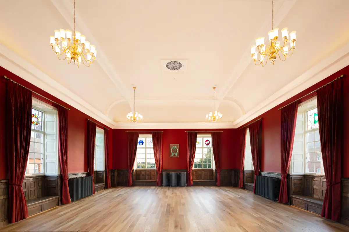 The council chamber, red walls and oak panelling under a coved ceiling, two brass chandeliers hanging over the empty floor and stained glass in the window heads