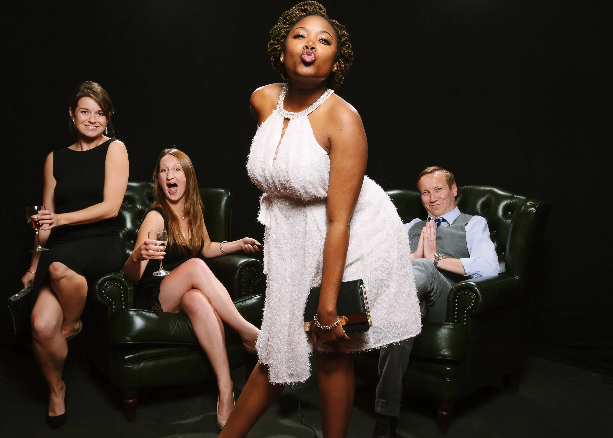 Four guests in evening dress on dark green leather armchairs against a black backdrop, one standing in front of them blowing a kiss to the camera