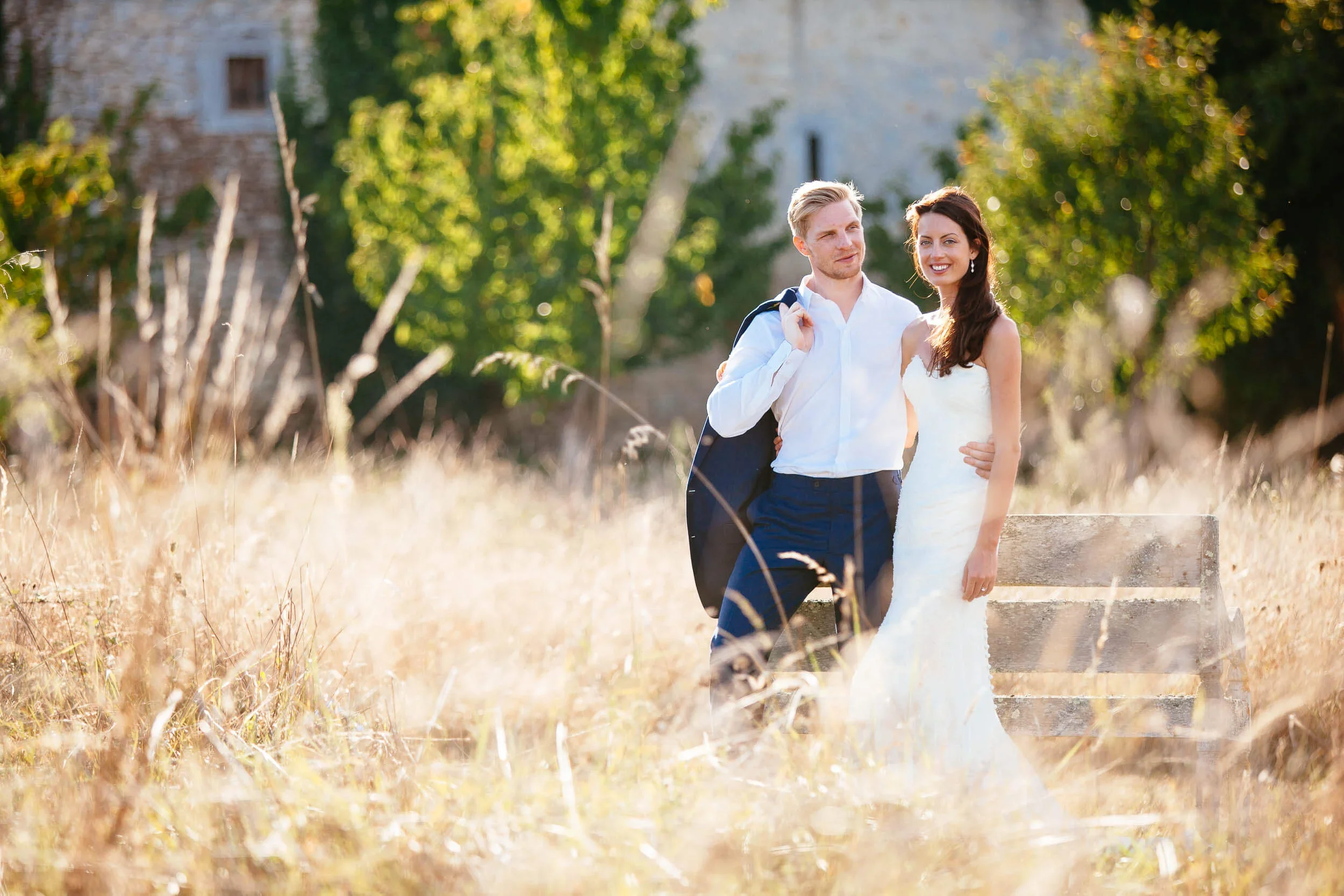 A bride and groom standing in long dry grass in low evening sun, a stone farmhouse behind them